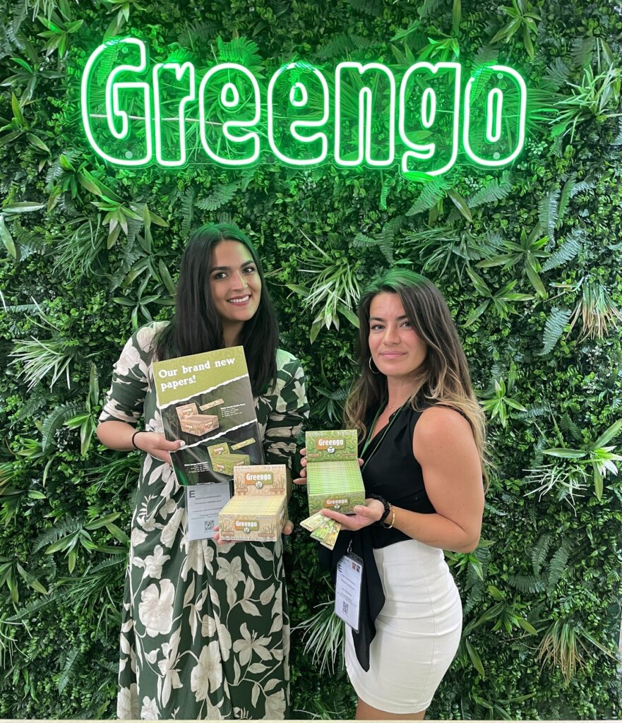 Greengo-stand op InterTabac met groene branding, felle lichten en twee vrouwen die Greengo-vloei producten vasthouden.